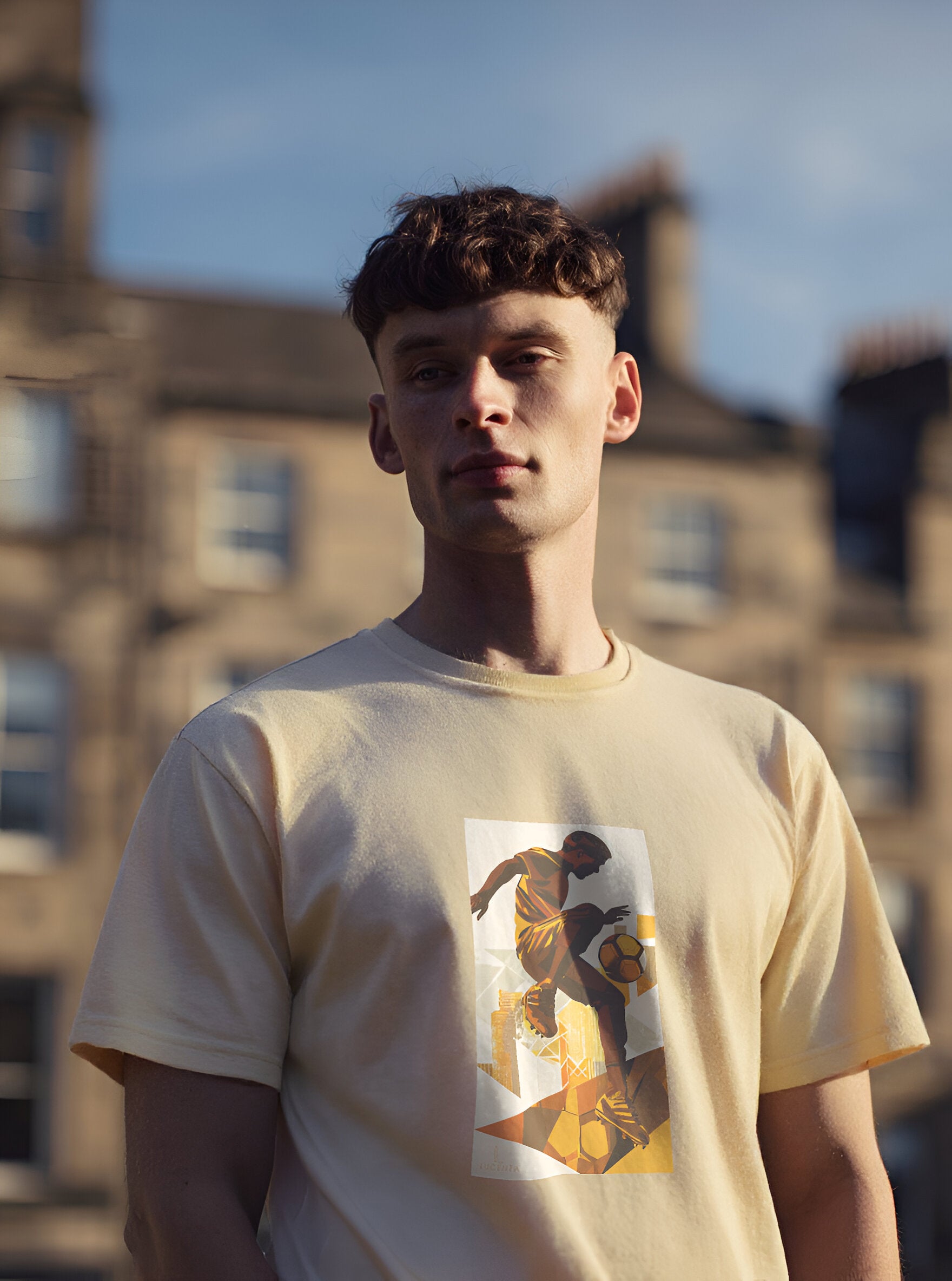 Man wearing a beige lucenza t-shirt with a graphic design, standing outdoors with buildings in the background.